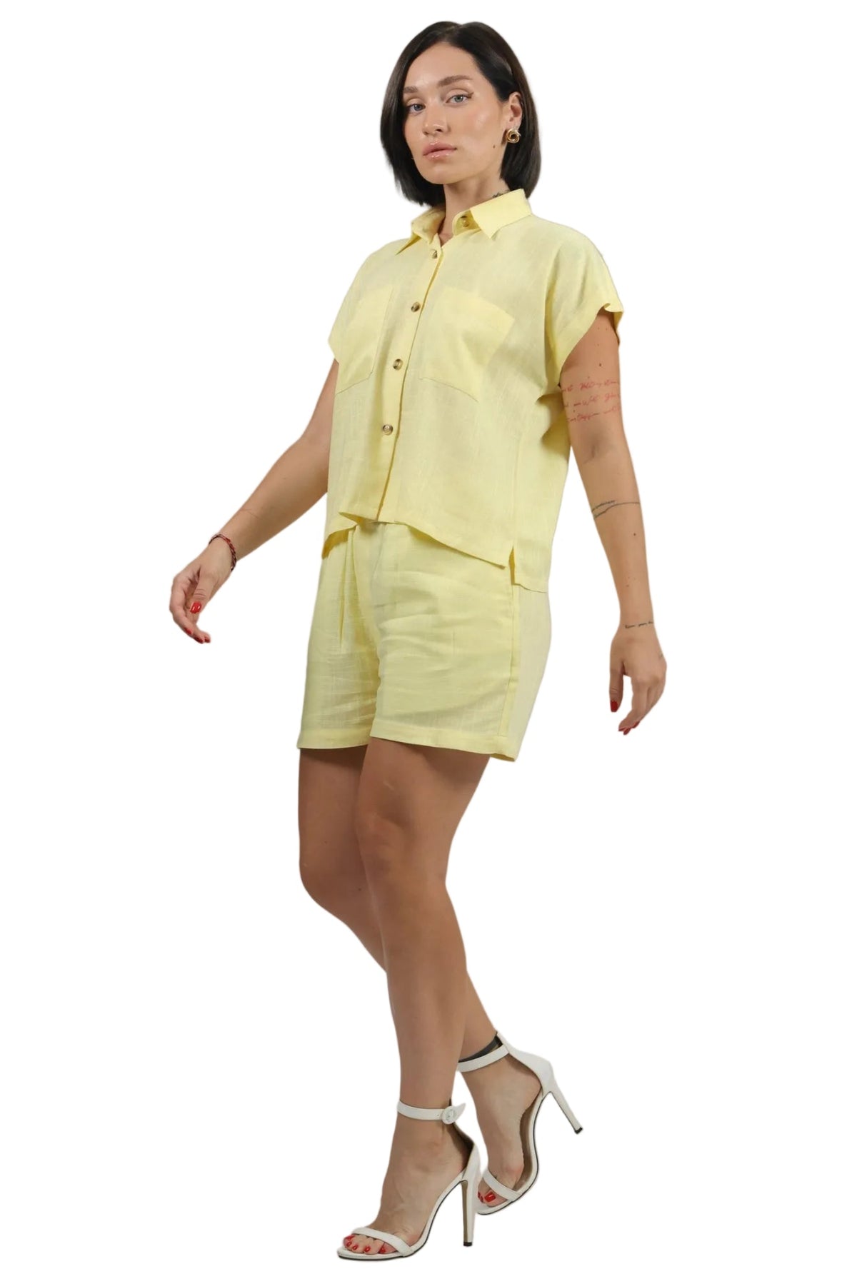 A woman modeling a light yellow short-sleeve button-up shirt and matching shorts set, paired with white high-heeled sandals, against a plain white background.