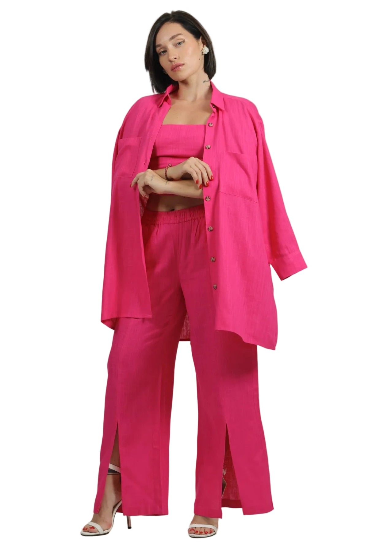 A woman modeling a bright pink matching outfit consisting of a long button-front overshirt, a fitted crop top, wide-leg trousers with front slits, and heeled sandals, photographed against a plain white studio background.