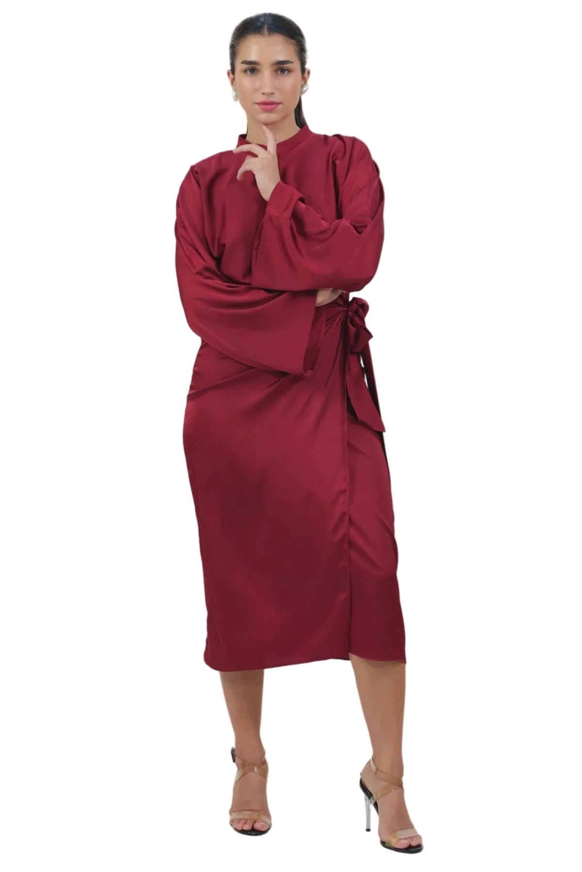 A woman modeling a long-sleeved, wrap-style midi dress in deep burgundy with a tie waist, paired with nude high-heeled sandals, against a solid white background.