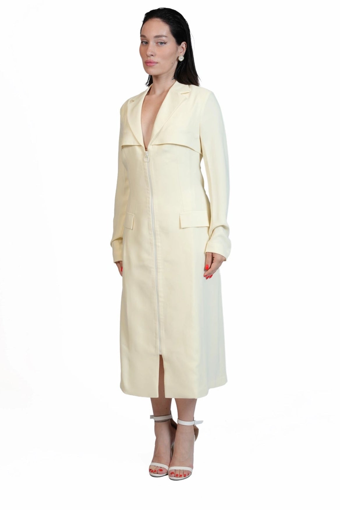 A woman modeling a long cream-colored trench coat with a deep V-neckline, wide lapels, and front pockets, paired with clear high-heeled sandals, set against a plain white background.