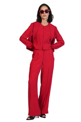A woman modeling a bright red two-piece outfit consisting of a long-sleeve cropped jacket with pleated shoulders and matching wide-leg trousers, paired with dark sunglasses and black heels, on a solid white background.