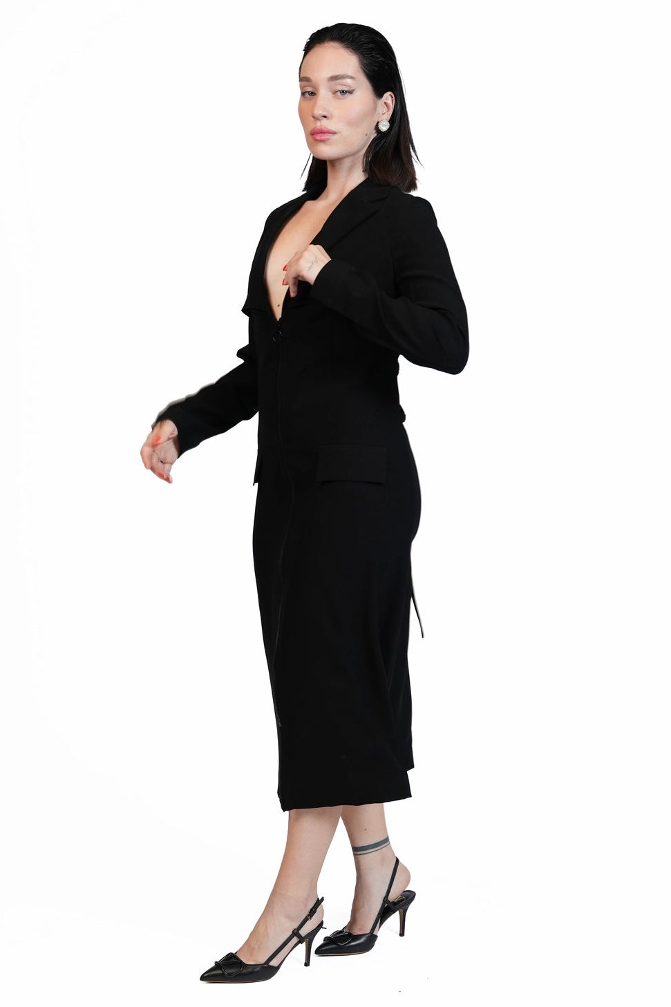 A woman modeling a black two-piece outfit consisting of a long-sleeve blazer with a deep V-neck and a matching knee-length skirt, paired with black pointed-toe heels, against a solid white background.