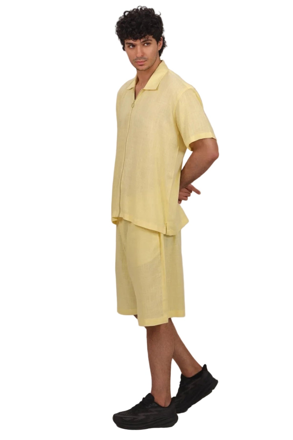 A man modeling a light yellow two-piece linen set consisting of a short-sleeve collared shirt and matching shorts, paired with black sneakers, against a solid white background.