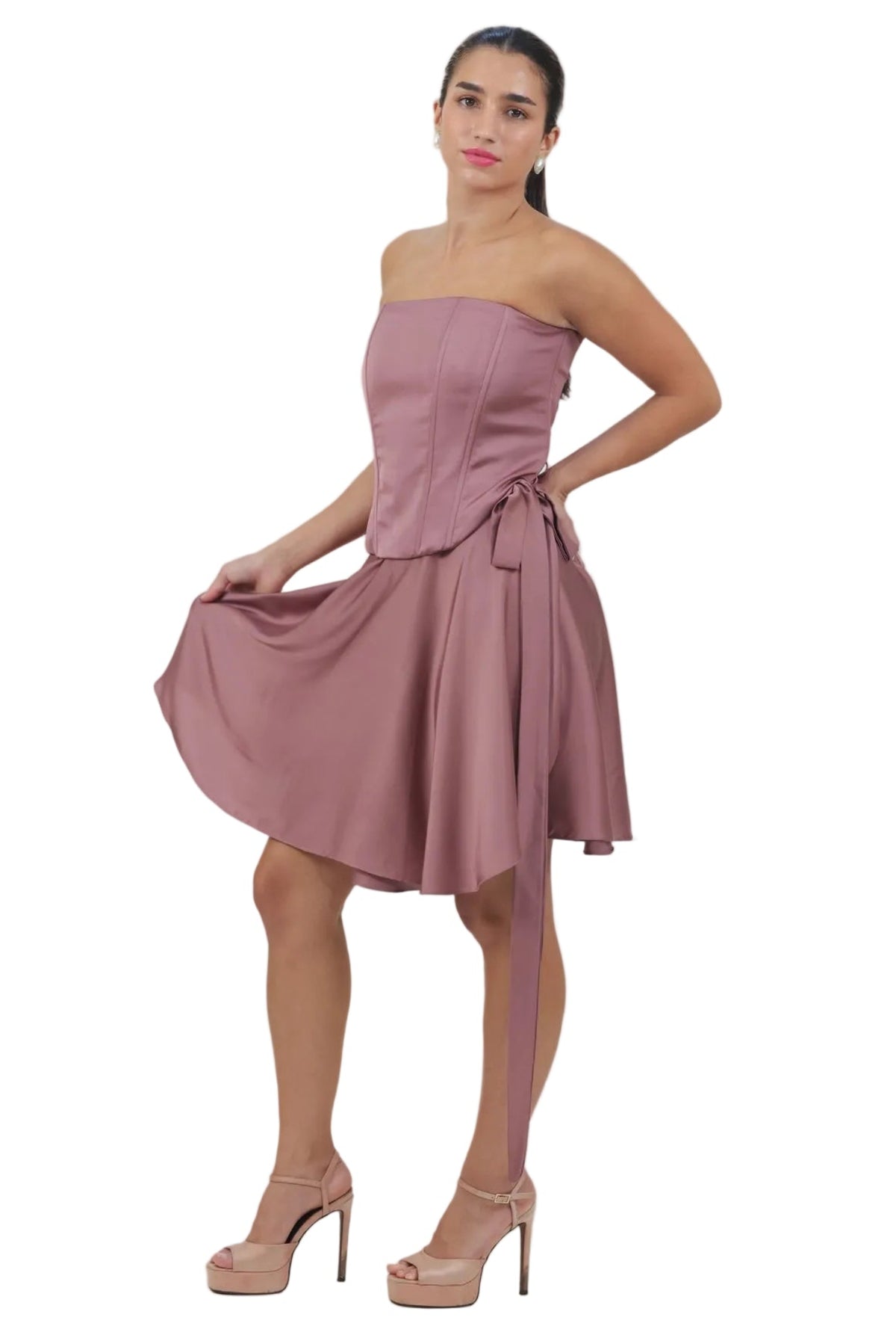 A woman modeling a strapless dusty rose mini dress with a corset-style bodice, flared skirt, and matching tie belt, paired with nude high heels against a plain white background.