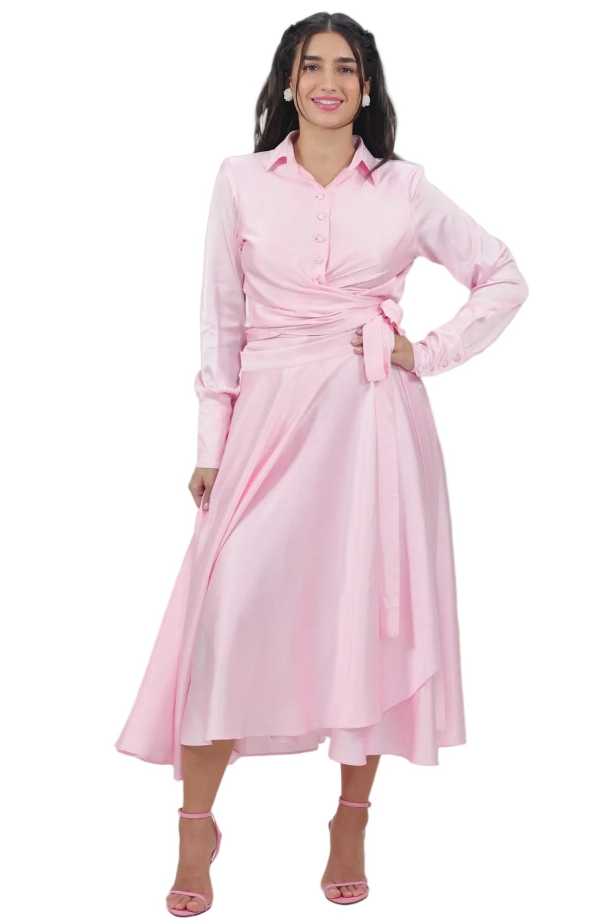 A woman modeling a light pink long-sleeve midi dress with a button-up collar, a wrap-style waist, and an asymmetrical draped skirt, paired with pink heeled sandals against a plain white background.