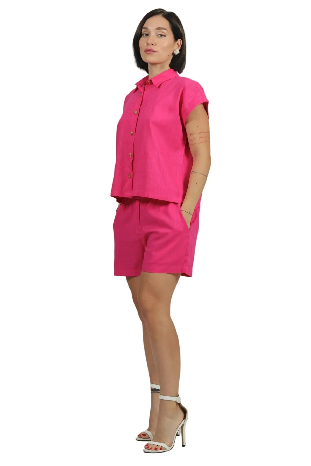 A woman modeling a bright pink two-piece set consisting of a short-sleeve button-up shirt and matching shorts, paired with white high-heeled sandals, against a plain white background.