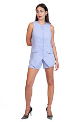 A woman modeling a light blue sleeveless romper with a button front and asymmetrical hem, paired with black high heels, on a solid white background.