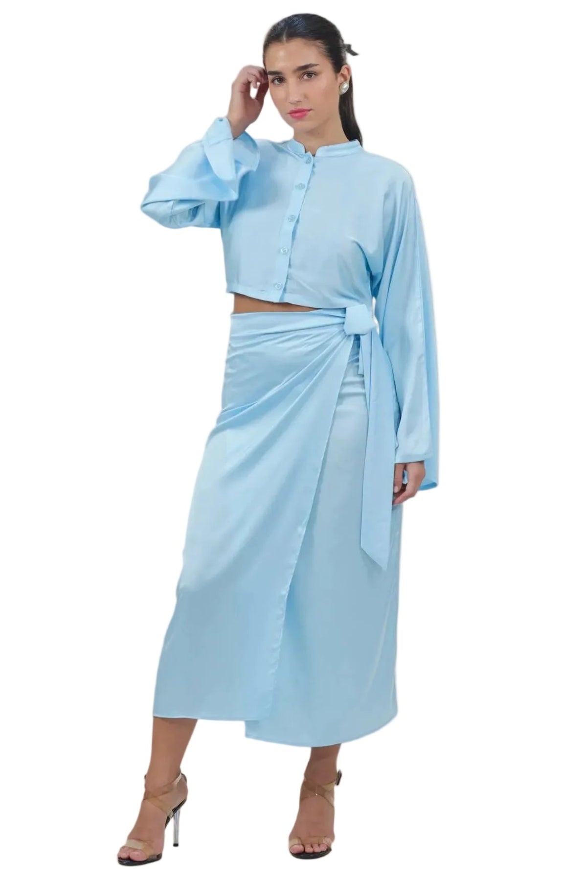 A woman modeling a light blue two-piece outfit consisting of a long-sleeve cropped button-front top and a matching high-waisted wrap midi skirt, shown against a plain white background.
