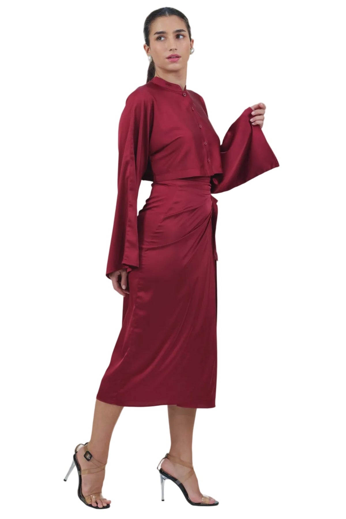 A woman modeling a long-sleeved, high-neck wrap-style midi dress in a deep burgundy red satin fabric, paired with clear high-heeled sandals, against a solid white background.