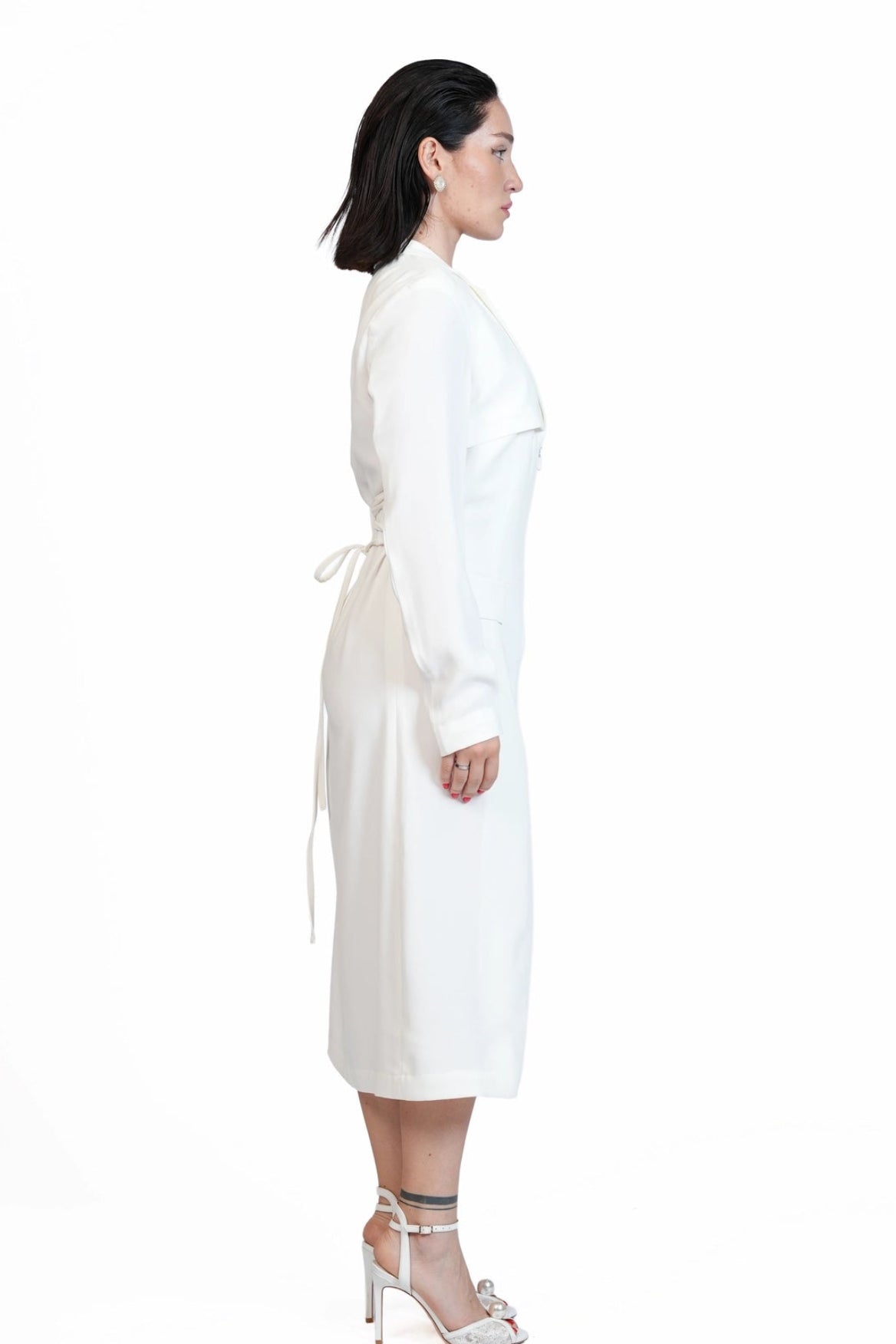 A woman shown in profile wearing a long-sleeved white wrap-style dress with a tie waist, paired with white high-heeled sandals, against a plain white studio background.