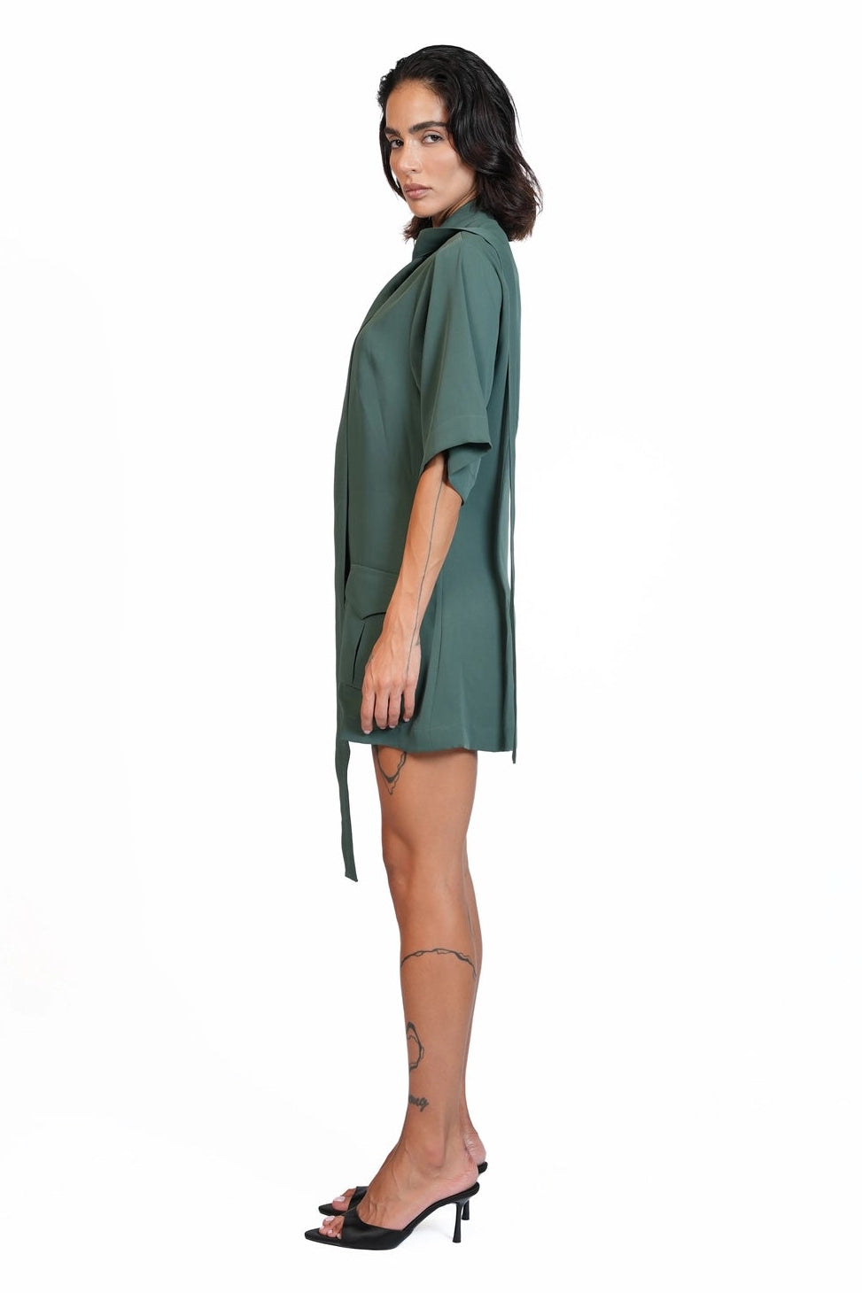 A woman modeling a sage green short-sleeve wrap dress with side ties, shown in profile against a solid white background.