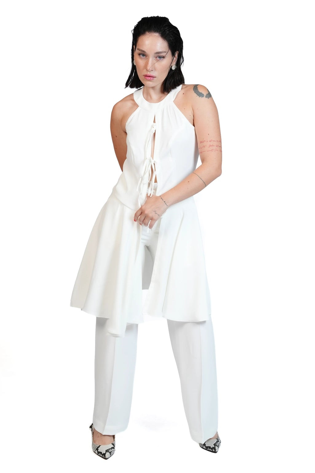 A woman modeling a white sleeveless halter top with a tie-front detail and a flared skirt, paired with matching white wide-leg pants and black pointed heels, against a solid white background.