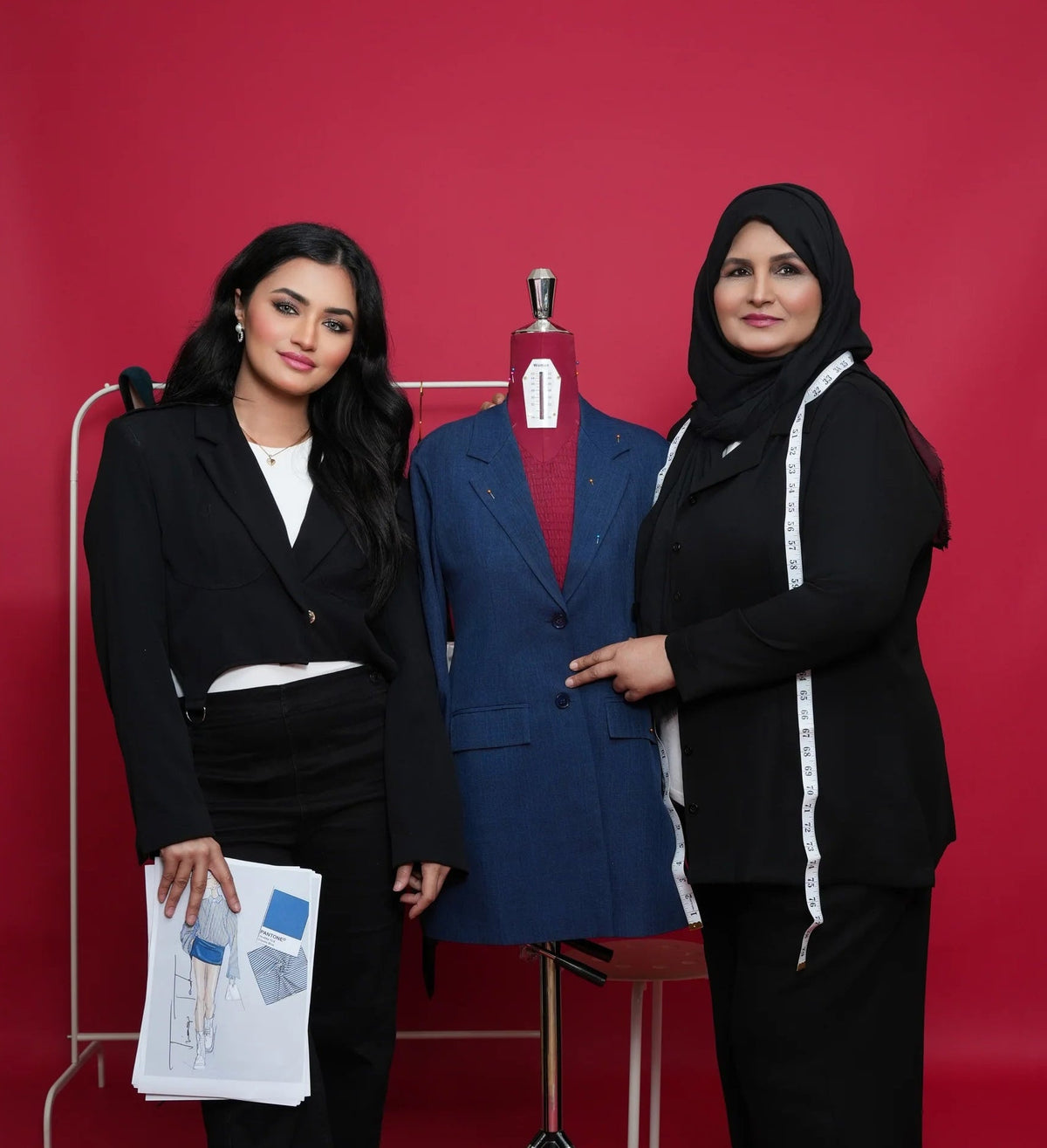 Two women standing next to a mannequin wearing a blue suit against a red background