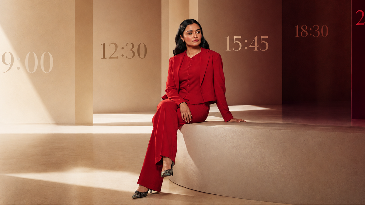A woman in a red long-sleeve top and matching wide-leg trousers sits on a curved beige platform in a minimalist interior with warm beige walls and large time numerals in the background.